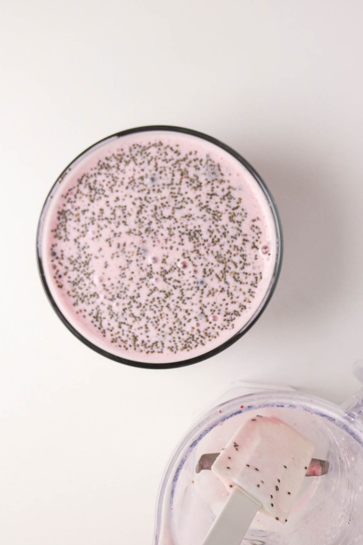 A bowl filled with pink chia seed pudding, viewed from above. Next to it is a blending container with a white spatula coated in the same pink mixture. Both are set on a white surface.