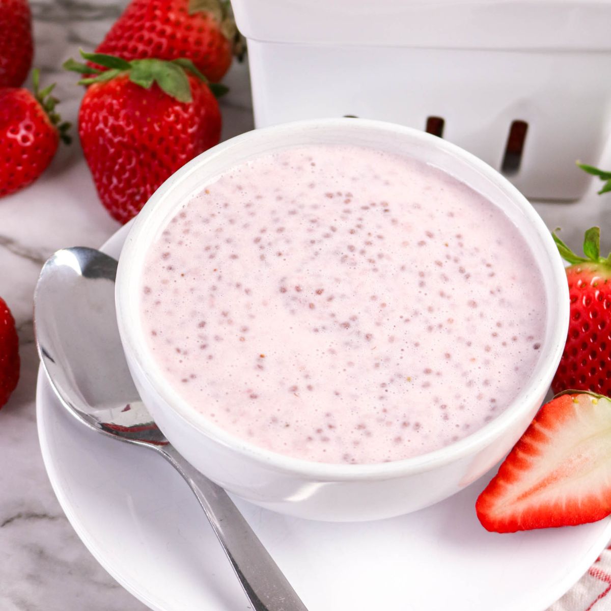 A white bowl filled with pink chia pudding sits on a white plate with a metal spoon. Whole and sliced strawberries are arranged around the bowl on a marble surface.