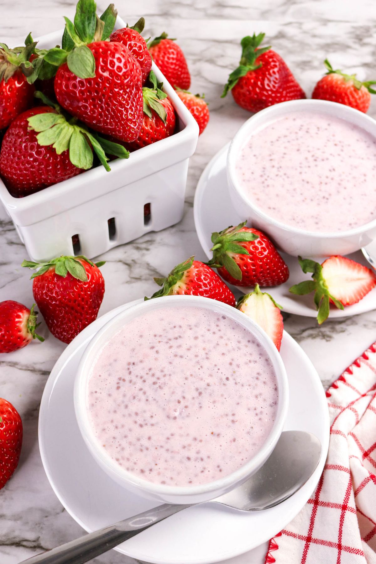 Two white bowls filled with pink chia seed pudding sit on white plates with spoons, surrounded by fresh strawberries and a white basket of strawberries on a marble surface. A red and white napkin is partially visible.
