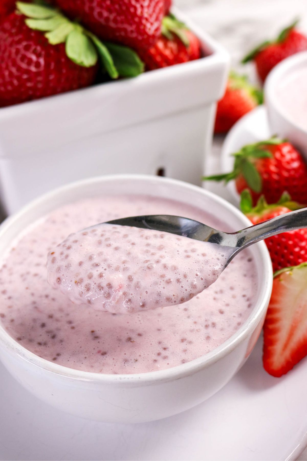 A spoonful of pink chia pudding is held above a small white bowl filled with more pudding. Fresh strawberries and a white container of strawberries are visible in the background.