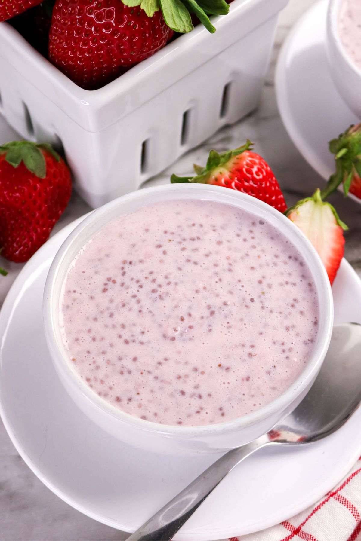 A white bowl filled with pink chia pudding sits on a white plate with a metal spoon. Fresh strawberries are placed around the bowl and a white container holding strawberries is in the background.