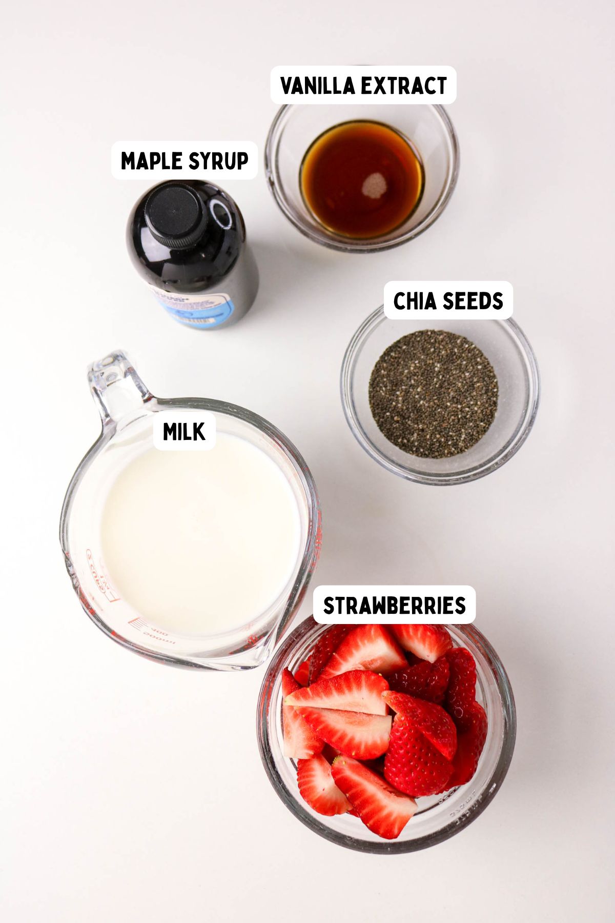 Five ingredients on a white surface: a bowl of vanilla extract, a bottle of maple syrup, a bowl of chia seeds, a measuring cup of milk, and a bowl of sliced strawberries, each labeled with text.