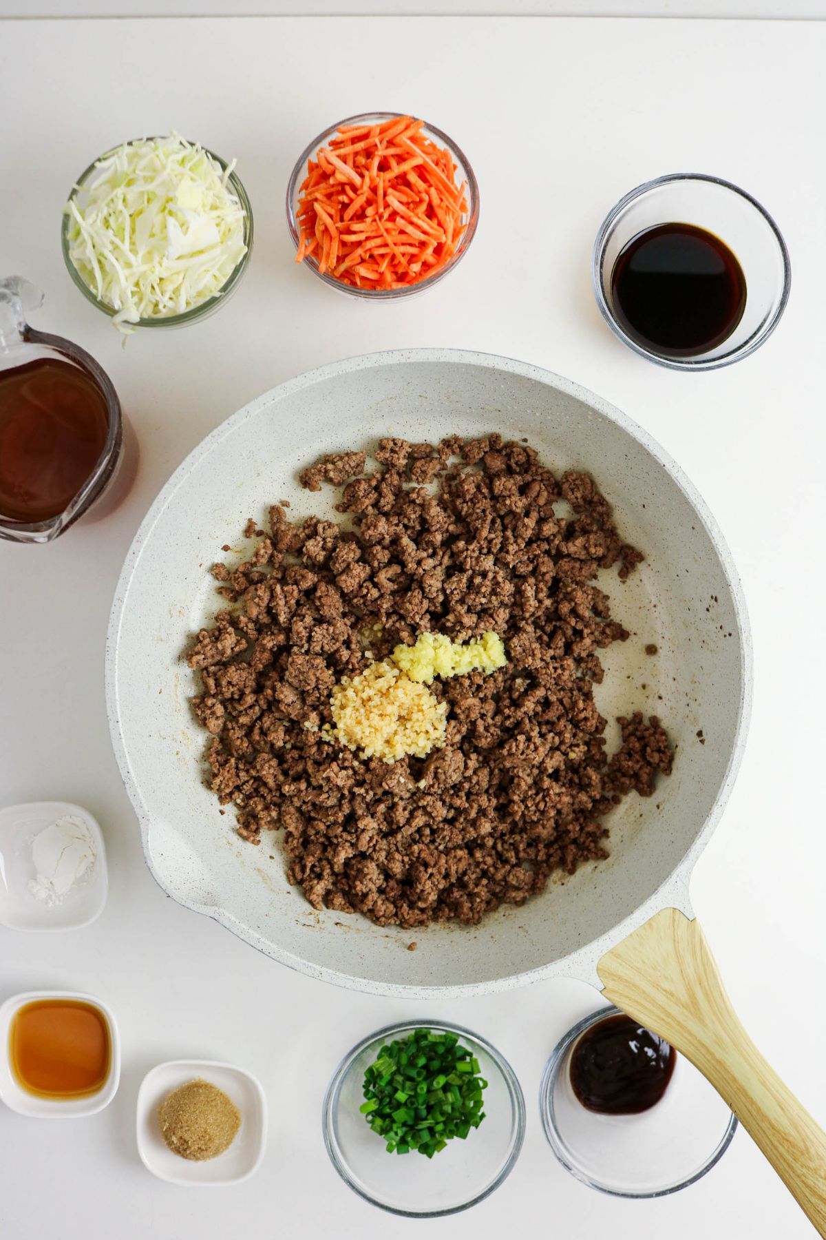 A white skillet with cooked ground beef and minced garlic, surrounded by small bowls containing shredded cabbage, shredded carrots, green onions, sauces, and spices on a white countertop.