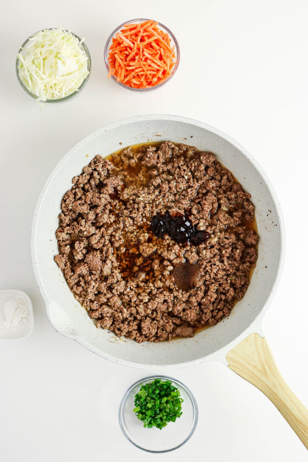 A white skillet with cooked ground beef and sauce in the center, surrounded by small bowls of shredded cabbage, shredded carrots, chopped green onions, and cornstarch on a white surface.