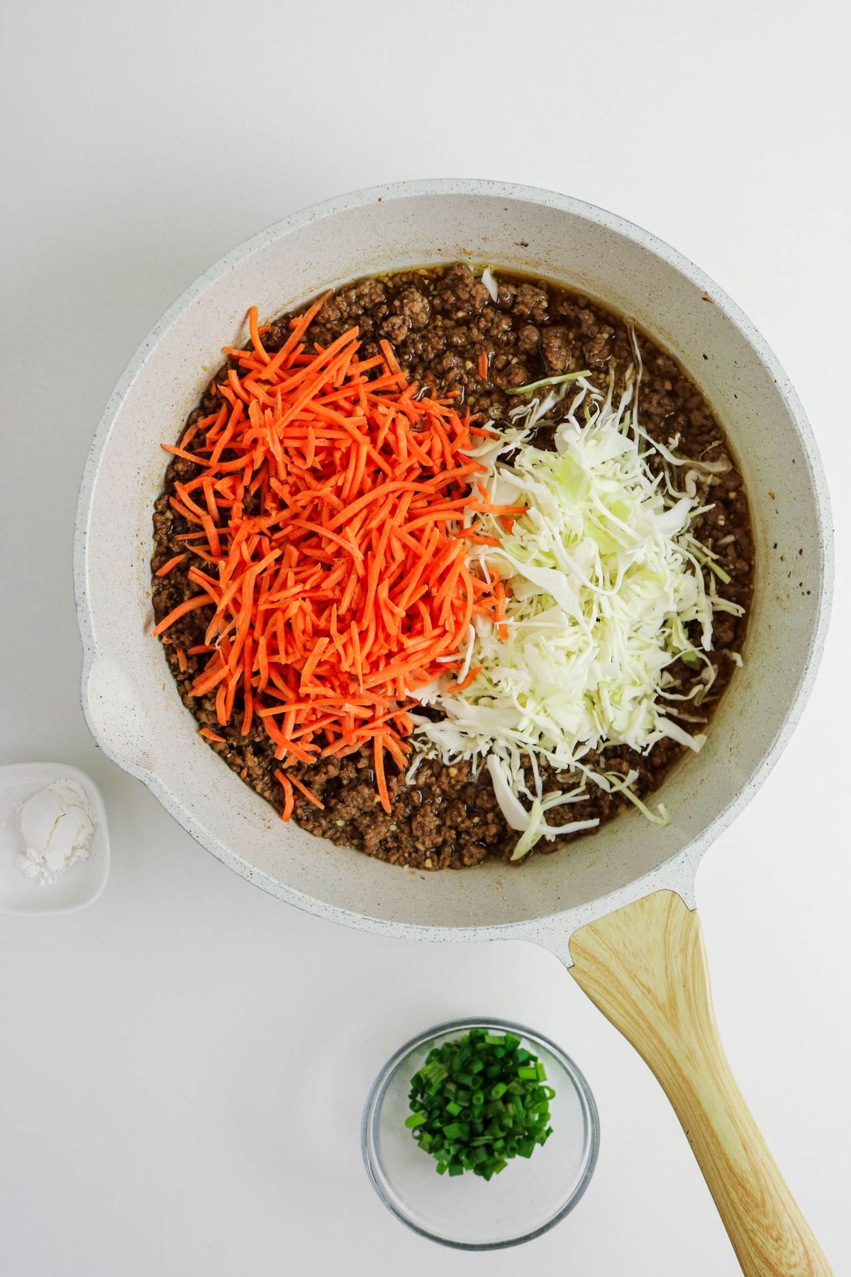 A white skillet with cooked ground beef, topped with piles of shredded carrots and shredded cabbage. A small bowl of chopped green onions and a small dish of white powder sit nearby on a white surface.