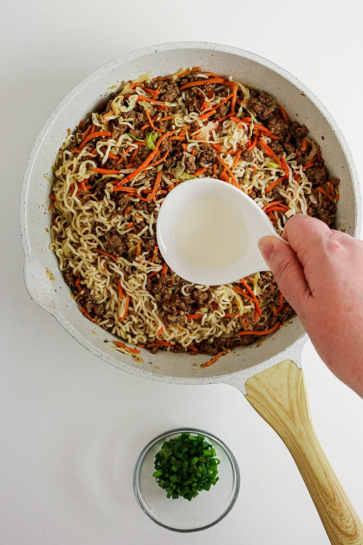A hand pours liquid from a small white bowl into a skillet containing cooked noodles, ground meat, shredded carrots, and cabbage. A small glass bowl with chopped green onions sits nearby on a white surface.