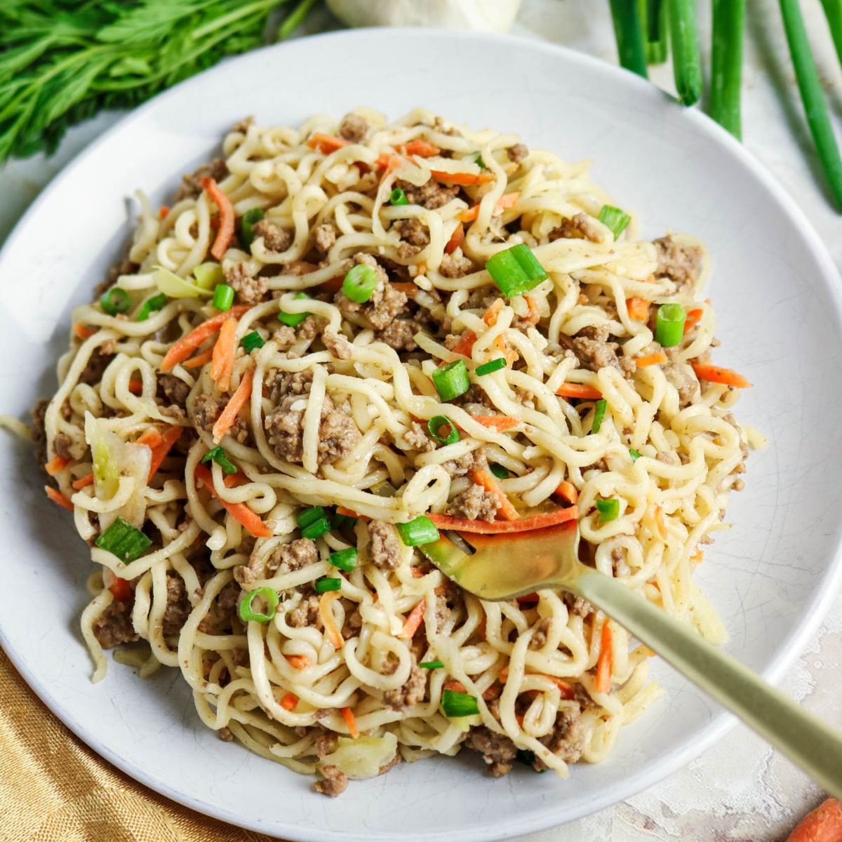 A plate of stir-fried noodles with ground meat, shredded carrots, and chopped green onions, served with a fork on the side. Green onions and a carrot are visible in the background.
