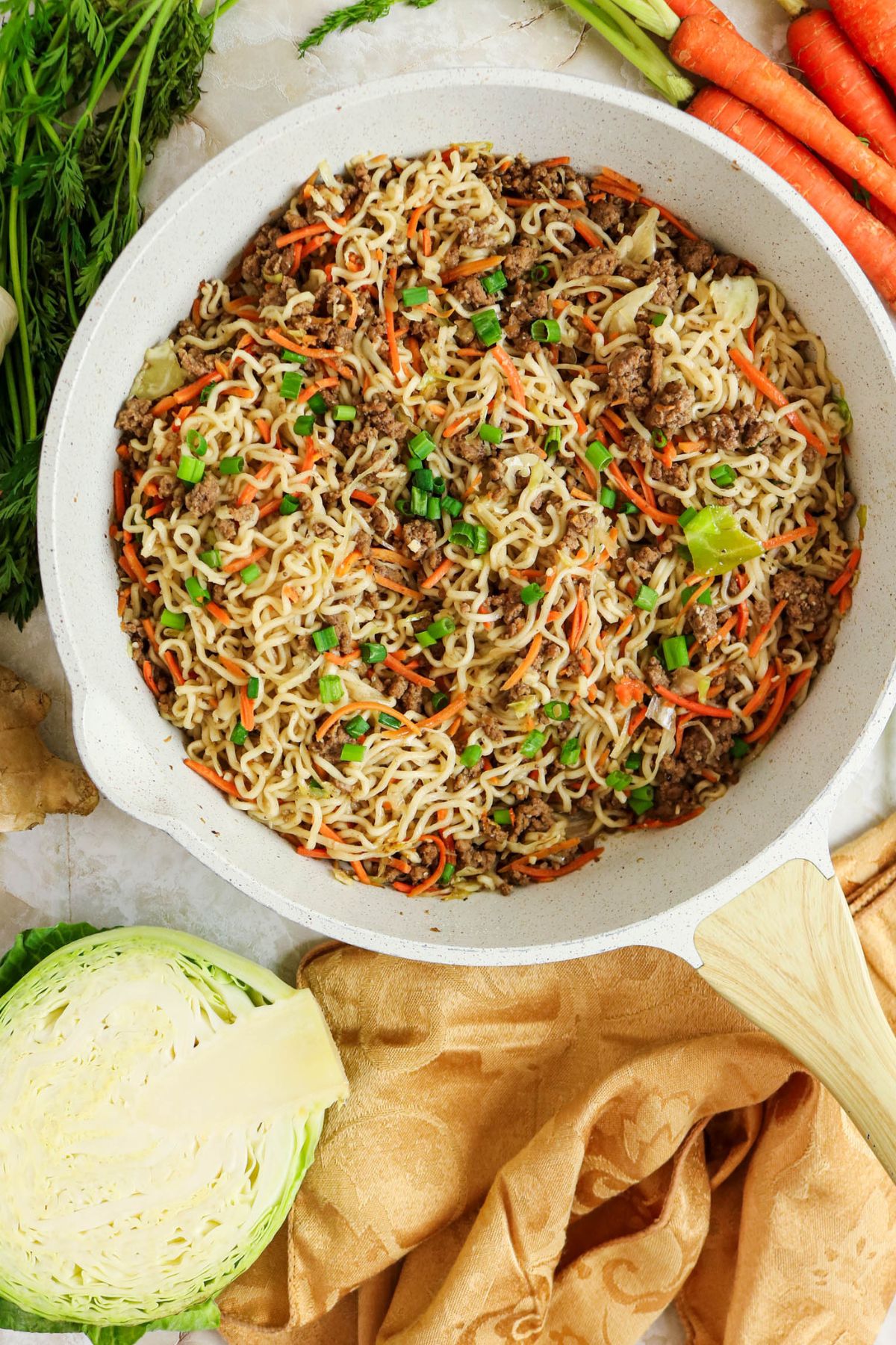 A white skillet filled with cooked noodles, ground beef, sliced carrots, cabbage, and green onions, surrounded by fresh carrots, cabbage, and a beige napkin. A wooden spatula rests on the skillet.