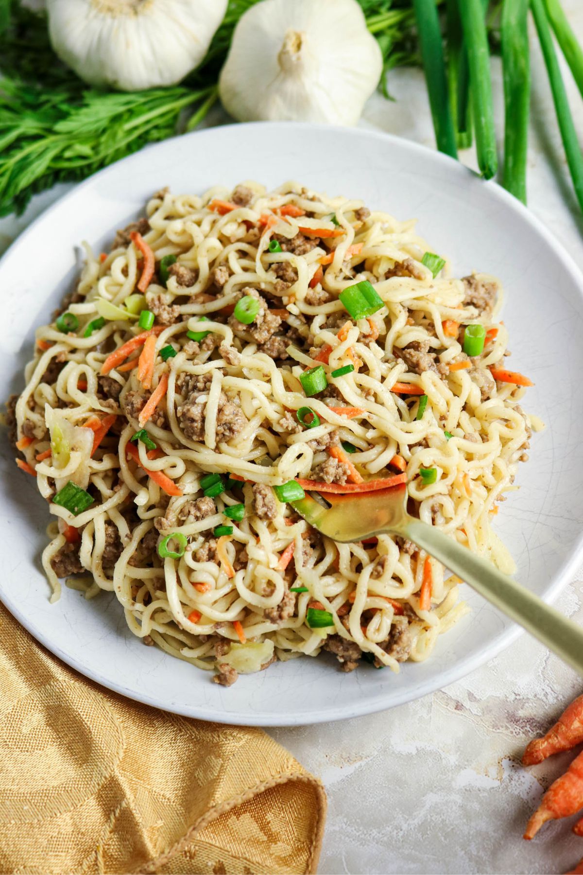 A plate of stir-fried noodles with ground meat, shredded carrots, and green onions, garnished with two heads of garlic and some fresh greens.