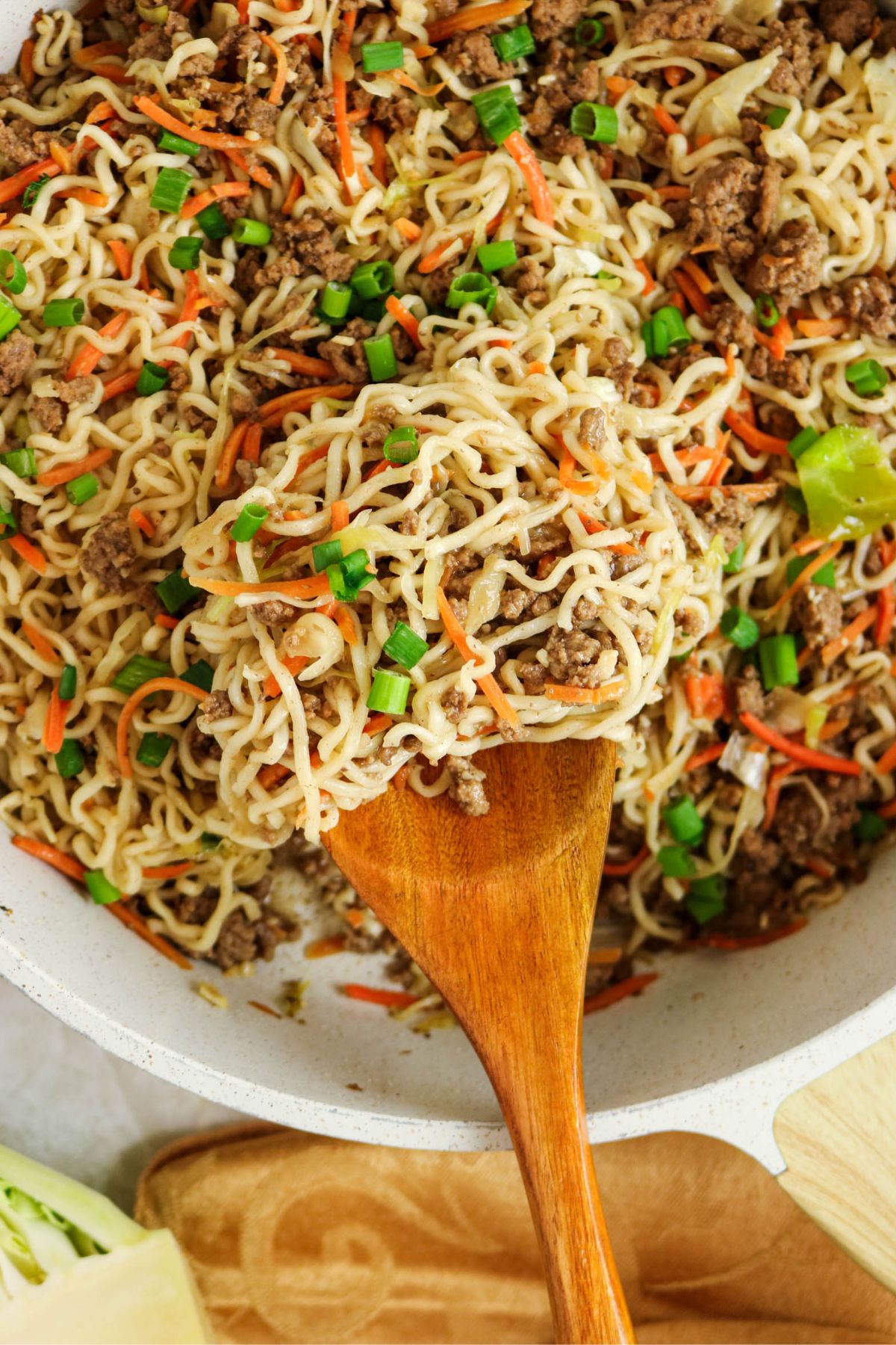 A close-up of cooked ramen noodles mixed with ground beef, shredded carrots, chopped green onions, and cabbage in a pan, with a wooden spoon lifting a portion.