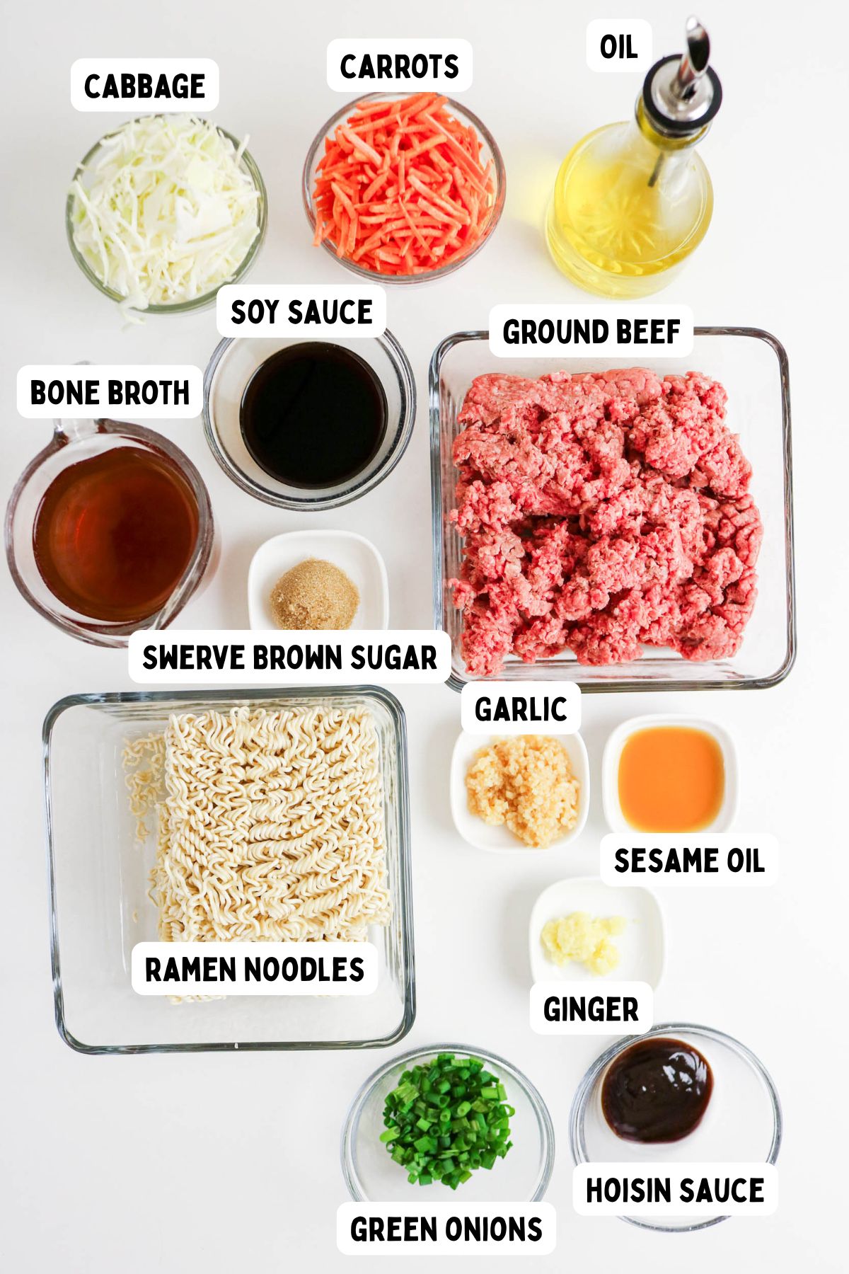 Top-down view of labeled ingredients for a noodle dish, including cabbage, carrots, oil, soy sauce, ground beef, bone broth, brown sugar substitute, ramen noodles, garlic, sesame oil, ginger, green onions, and hoisin sauce.
