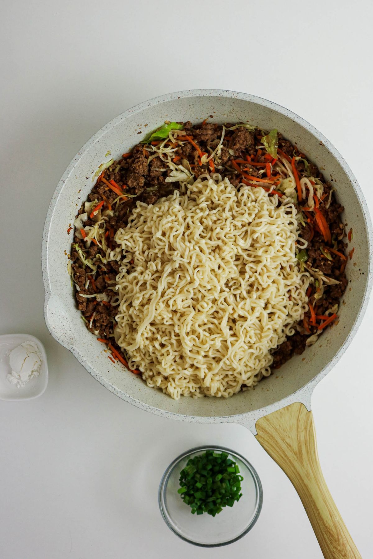 A white skillet containing cooked ground beef, shredded carrots, cabbage, and a pile of cooked ramen noodles on top. Nearby are a small bowl of chopped green onions and a small dish with a white powder, on a white surface.