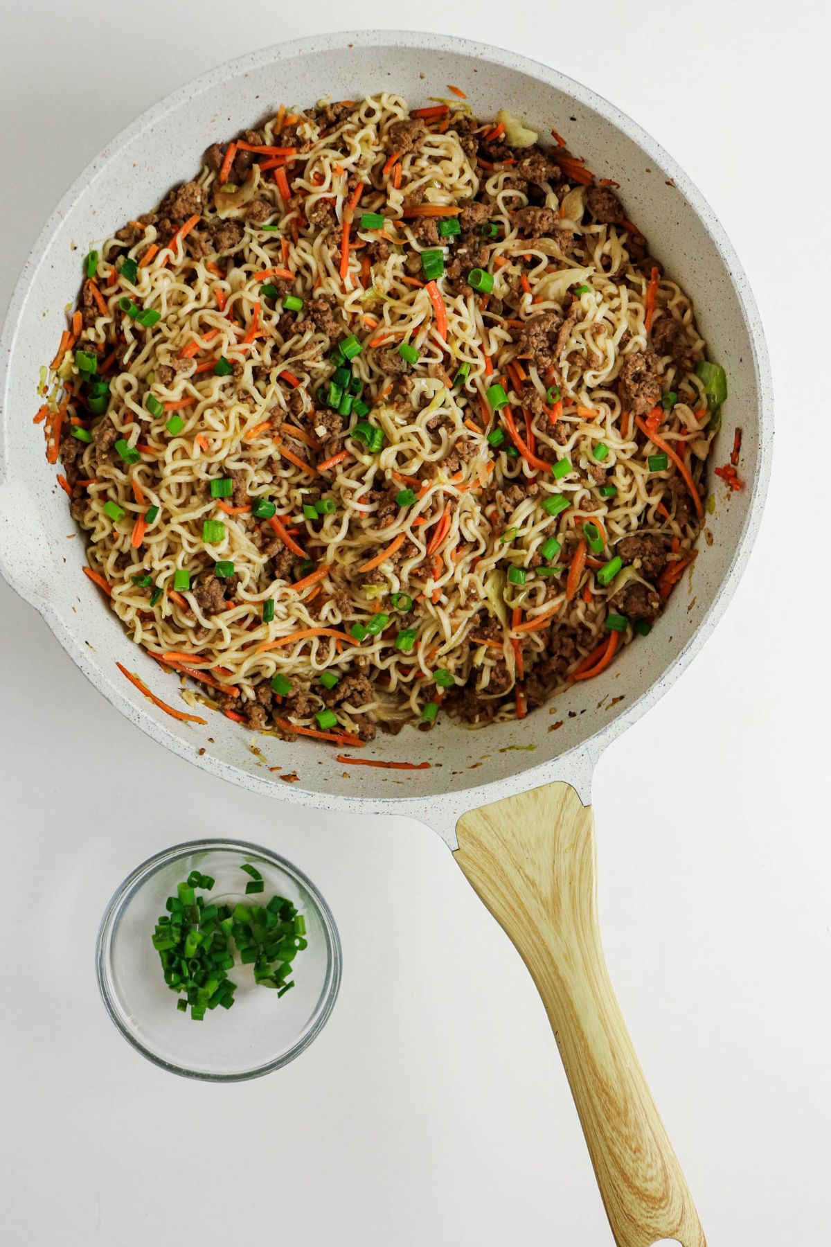A white skillet filled with cooked noodles, ground meat, shredded carrots, and green onions sits on a white surface. Beside it is a small clear bowl containing chopped green onions.