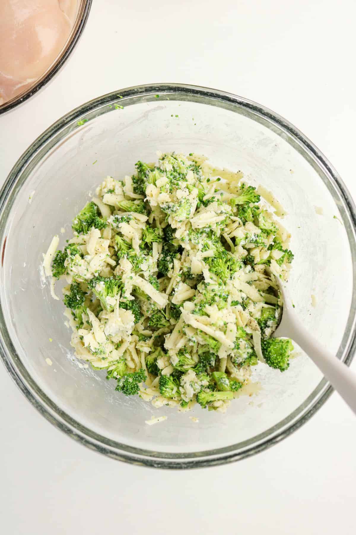A clear glass bowl contains a mixture of chopped broccoli, shredded cheese, and other finely chopped ingredients—perfect for preparing chicken and broccoli stuffed chicken. A white spoon rests in the bowl against a clean white background.