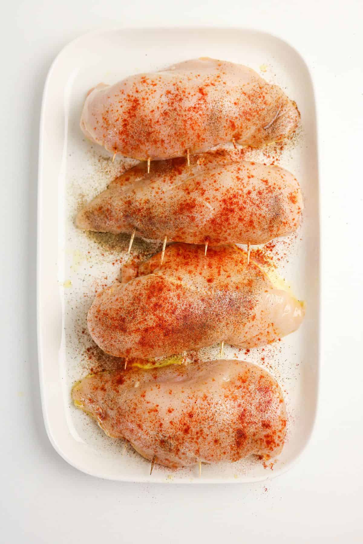 Four raw chicken breasts on a white rectangular plate, each secured with toothpicks and sprinkled with red seasoning and black pepper—perfect for making flavorful chicken and broccoli stuffed chicken.