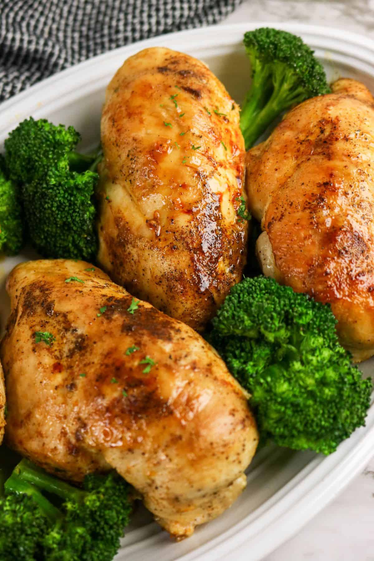 Three seasoned, cooked chicken breasts are arranged on a white plate with several pieces of steamed broccoli—creating a tempting twist on classic chicken and broccoli stuffed chicken. The golden-brown chicken glistens against a textured black-and-white cloth backdrop.