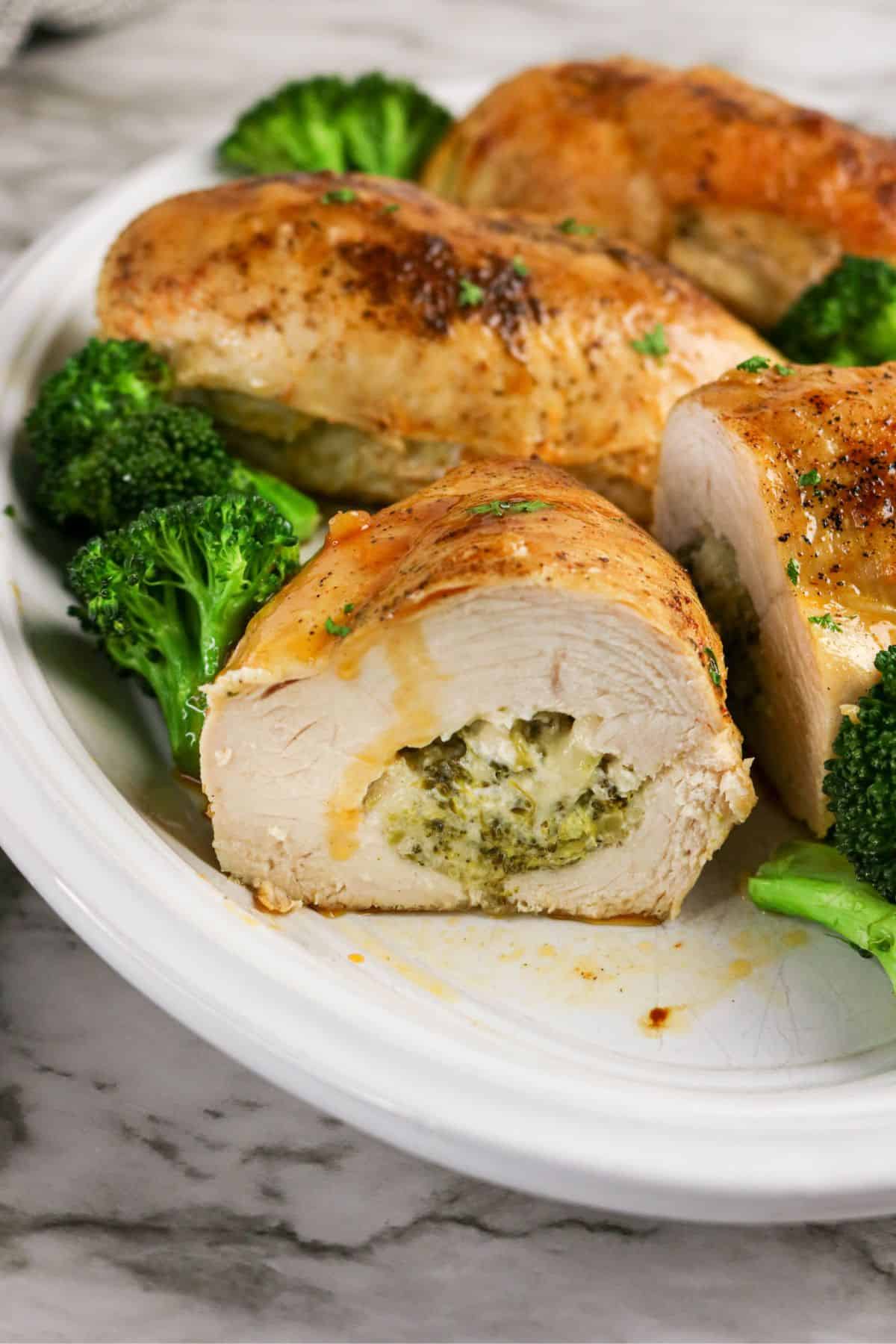 A sliced chicken and broccoli stuffed chicken breast on a white plate reveals a melted cheese and herb filling, surrounded by broccoli florets and whole chicken breasts. The plate rests on a marble countertop.