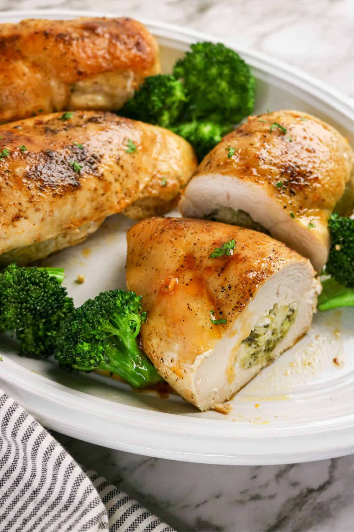 Sliced chicken and broccoli stuffed chicken breasts are filled with a creamy mixture, served alongside broccoli florets on a white oval plate. Garnished with herbs, the dish rests on a marble surface beside a striped napkin.