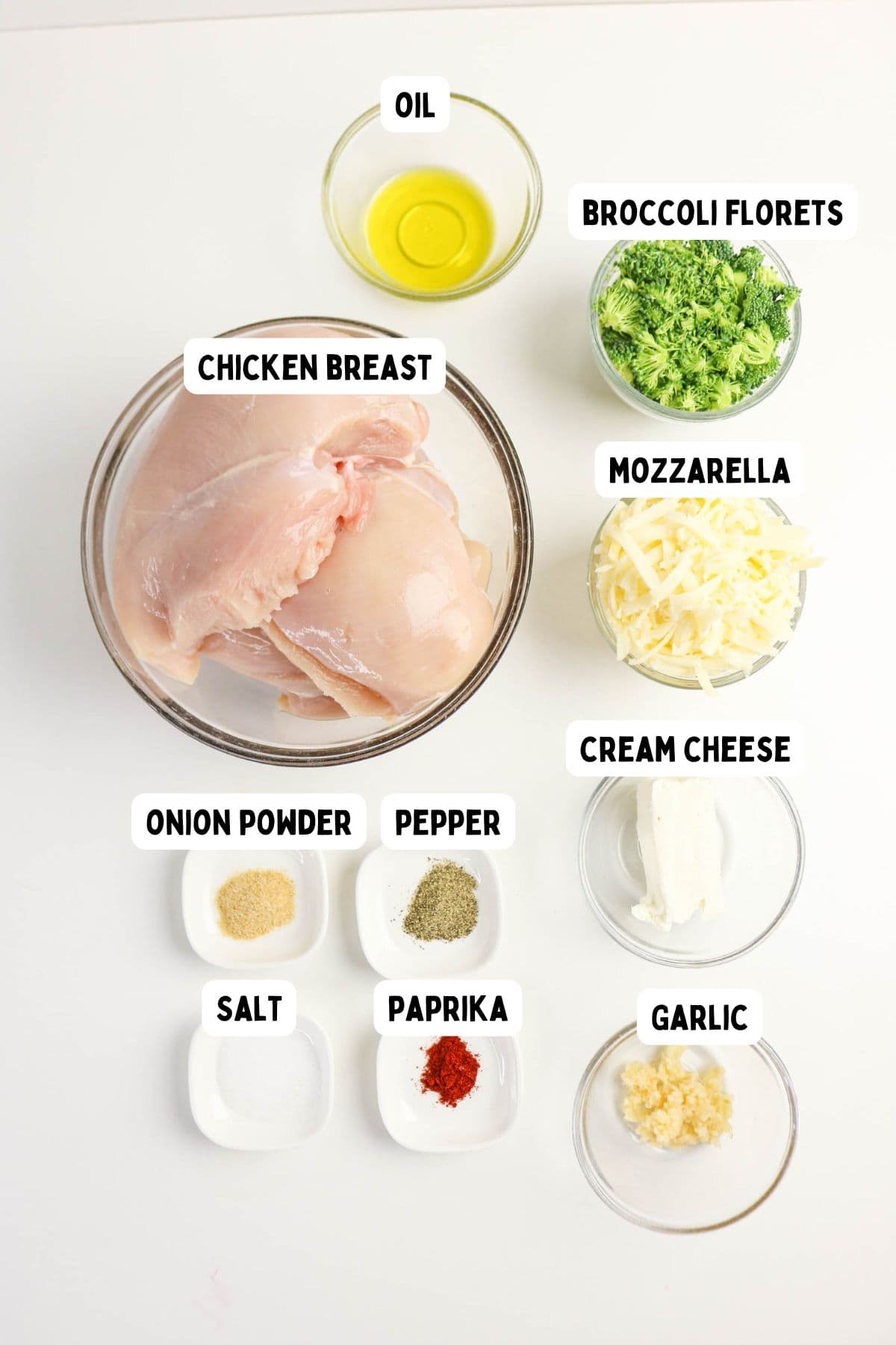 A top-down view of ingredients in bowls and dishes for chicken and broccoli stuffed chicken—oil, chicken breast, broccoli florets, mozzarella, cream cheese, garlic, paprika, pepper, onion powder, and salt—neatly arranged on a white surface.