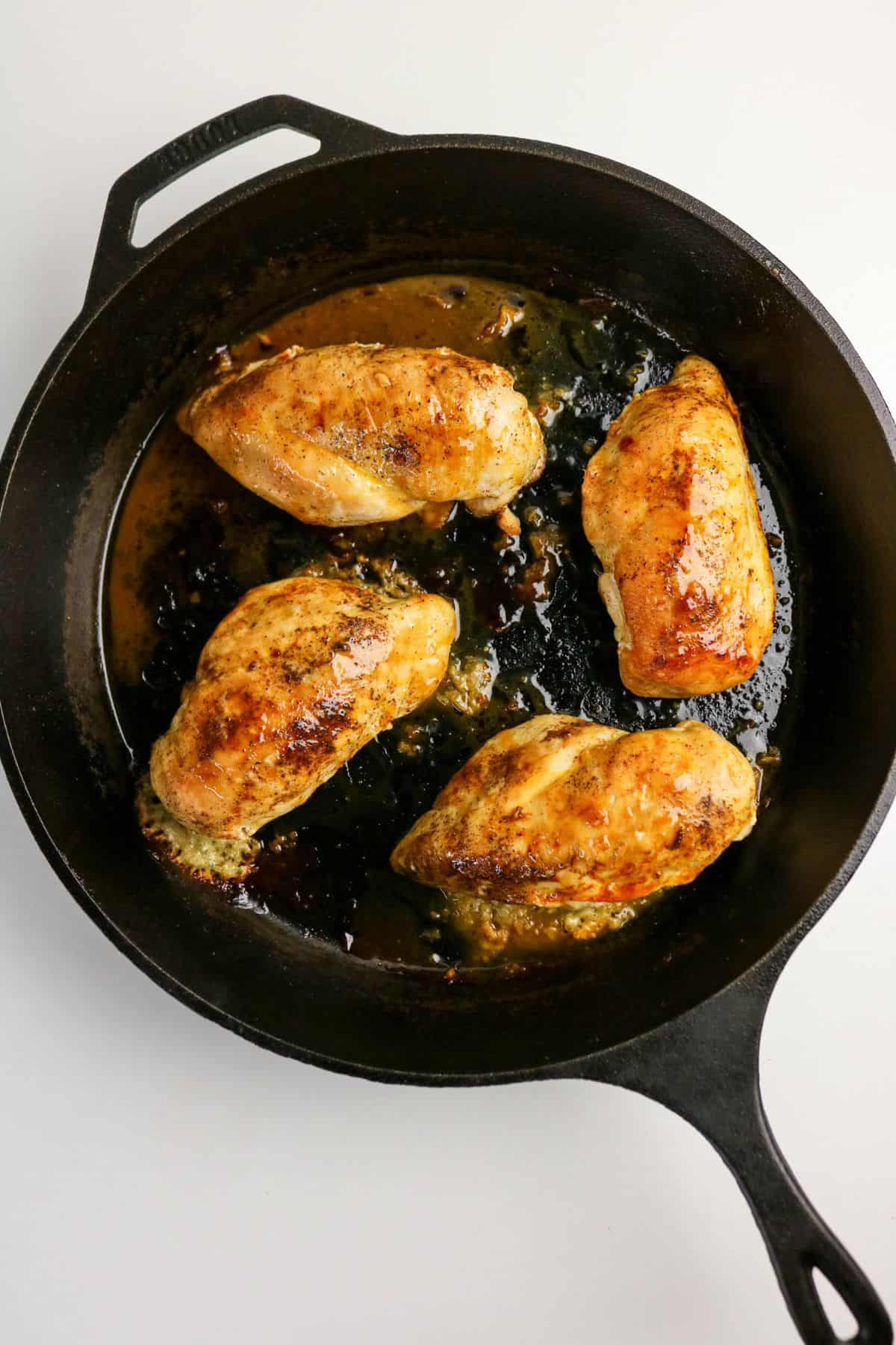 Four cooked, golden-brown chicken breasts are arranged in a cast iron skillet with some juices on the bottom, resembling a classic chicken and broccoli stuffed chicken dish. The skillet sits on a plain white surface.