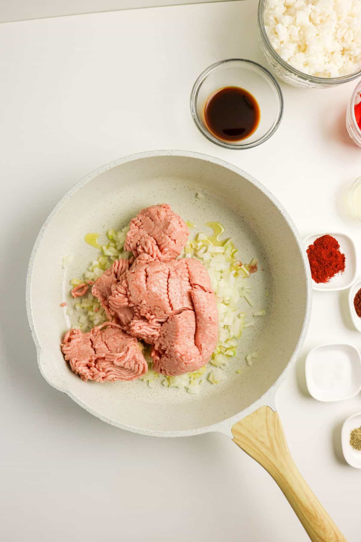 A white skillet with raw ground turkey and chopped onions on a white surface, surrounded by small bowls containing spices, soy sauce, rice, and other ingredients.