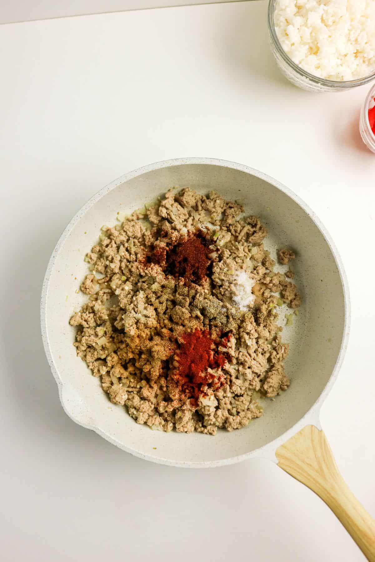 A white frying pan with cooked ground meat topped with red and brown spices sits on a white surface. Nearby, a glass bowl with white crumbled cheese and part of another bowl are visible.