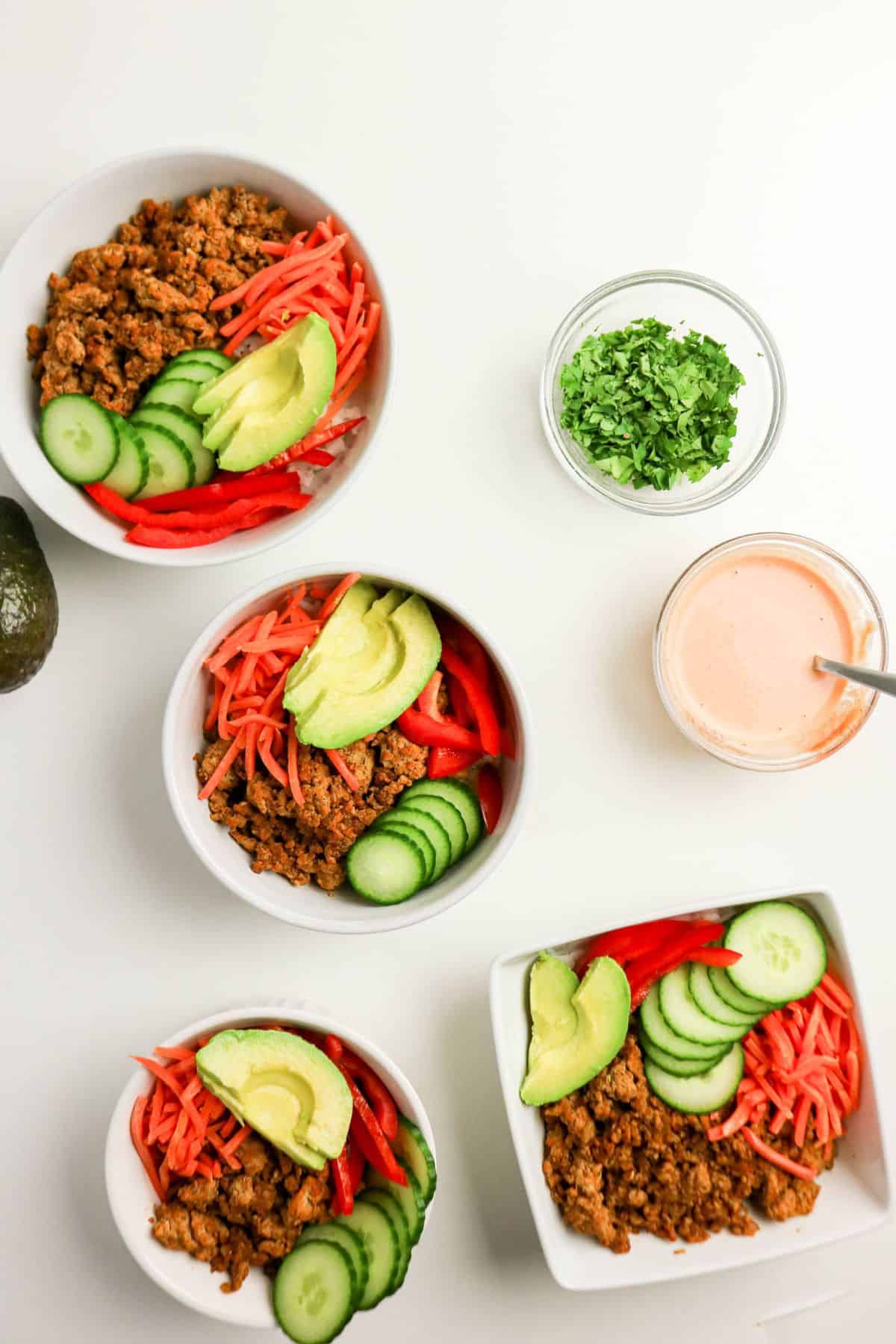 Four bowls filled with ground meat, sliced cucumbers, sliced avocado, shredded carrots, and red bell peppers on a white surface. A small bowl of chopped herbs and a jar of pink sauce with a spoon are nearby.
