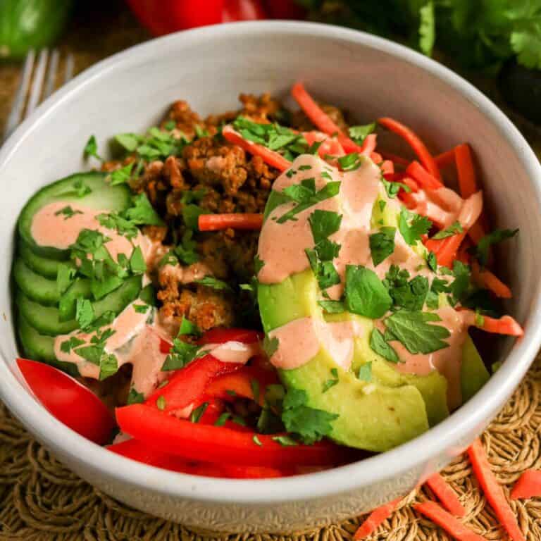 A white bowl filled with sliced cucumber, red bell pepper, shredded carrots, avocado, ground meat, and a pinkish dressing, topped with chopped fresh herbs. The bowl is placed on a woven mat.