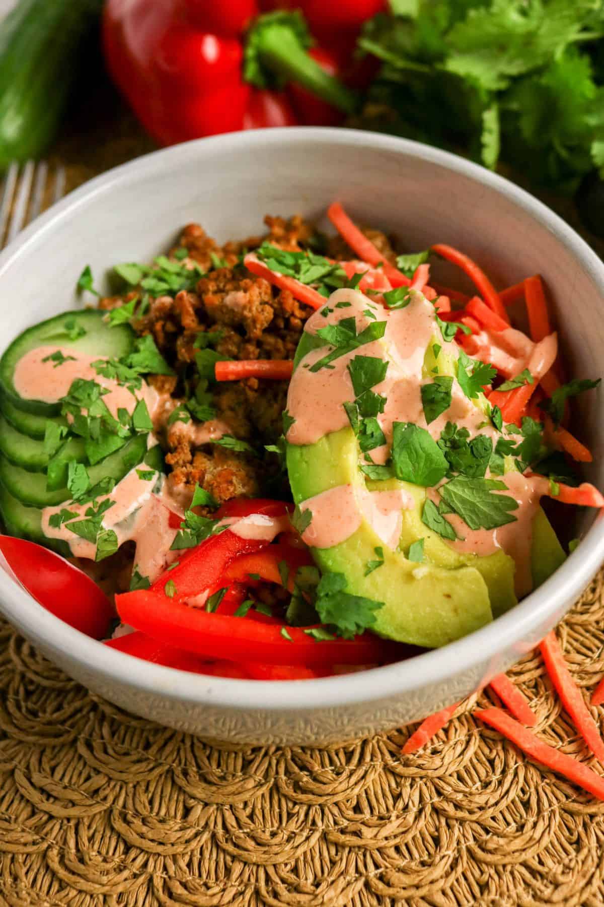 A bowl filled with sliced cucumber, avocado, red bell pepper, shredded carrots, ground meat, fresh cilantro, and a drizzle of creamy dressing, placed on a woven mat with vegetables in the background.