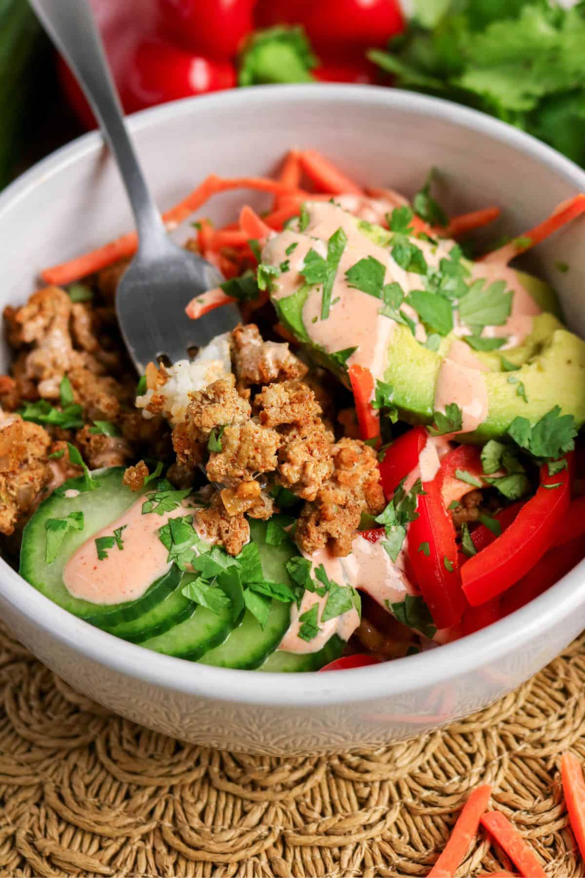 A bowl with seasoned ground meat, sliced cucumbers, avocado, red bell peppers, shredded carrots, chopped cilantro, and a drizzle of creamy sauce. A fork is placed in the bowl. Fresh vegetables are visible in the background.