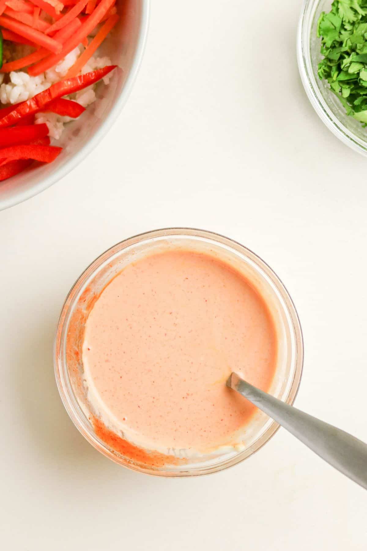 A small glass bowl filled with a creamy, light orange sauce sits on a white surface. A spoon rests in the bowl. Nearby are partial bowls of sliced vegetables and chopped green herbs.