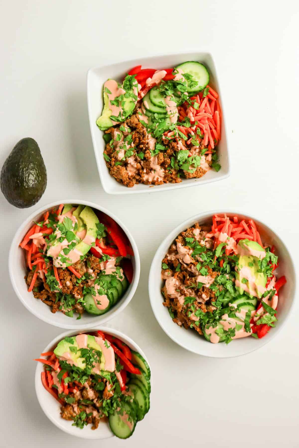 Four bowls containing a mixture of quinoa, sliced cucumber, red bell pepper, avocado, and chopped cilantro, topped with a light drizzle of sauce, are arranged on a white surface next to a whole avocado.