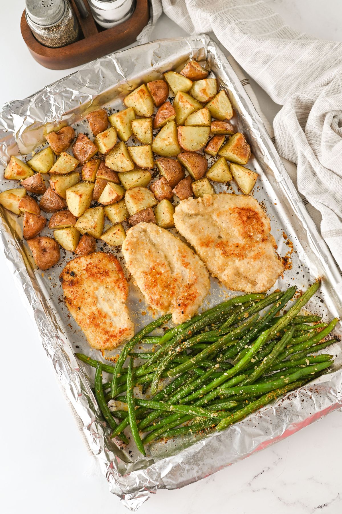 A foil-lined baking sheet with three baked parmesan crusted chicken breasts, roasted potato pieces, and a portion of seasoned green beans. A salt and pepper set and a beige cloth napkin are nearby.