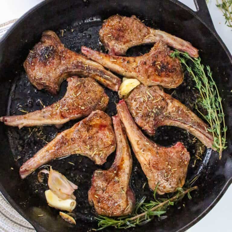 Six pan seared lamb chops in a cast iron skillet with sprigs of rosemary, thyme, and garlic cloves. The lamb chops are browned and seasoned, and the skillet is shown from above.