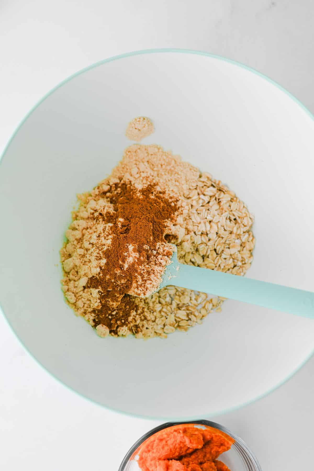 A white mixing bowl contains oats, brown sugar, and ground cinnamon for Pumpkin Overnight Oats, with a teal spatula inside. A small glass bowl with orange pumpkin puree is partially visible at the bottom.
