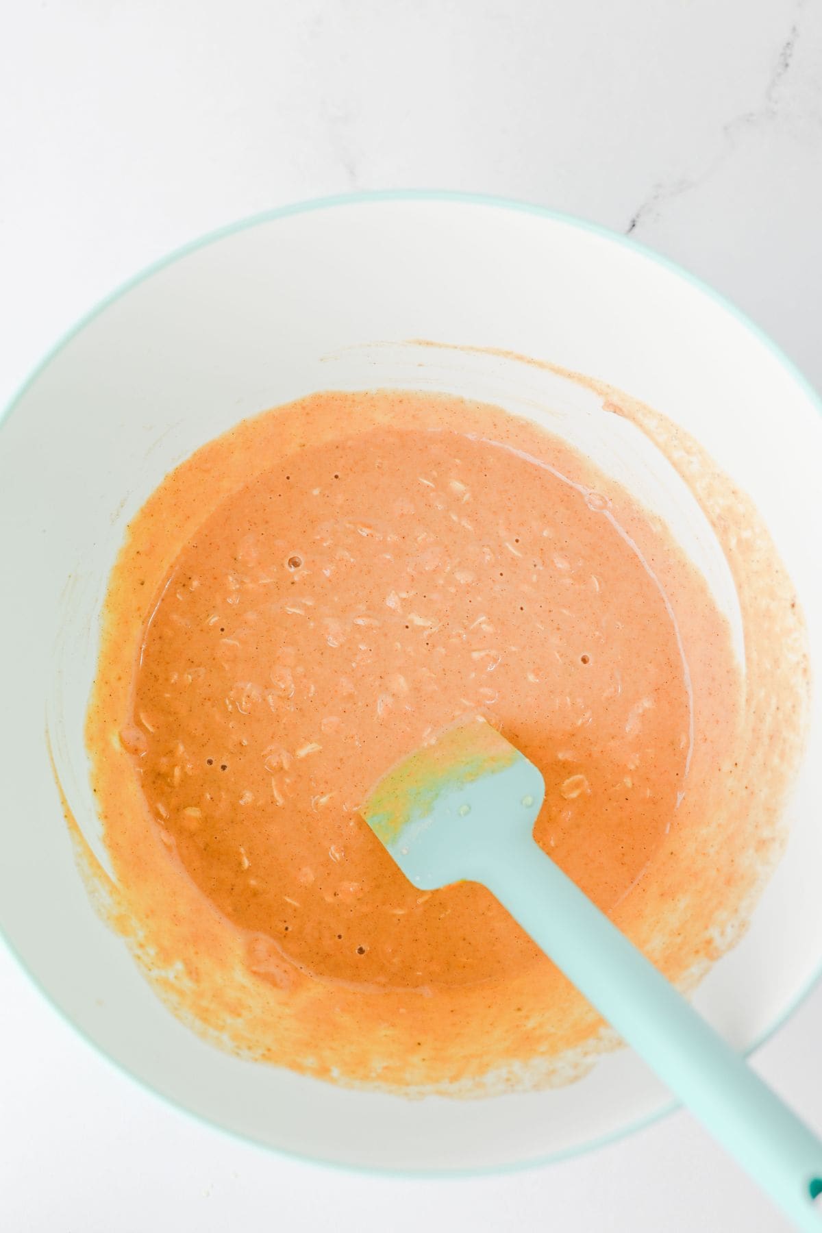 A light blue spatula rests in a white mixing bowl containing a light brown, slightly lumpy batter—reminiscent of the cozy texture found in Pumpkin Overnight Oats. The bowl sits on a white surface, and some batter is smeared along the sides of the bowl.