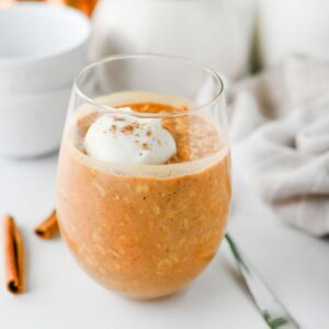 A clear glass filled with orange-brown Pumpkin Overnight Oats, topped with a dollop of whipped cream and a sprinkle of cinnamon. A cinnamon stick and a spoon are placed nearby on a white surface.