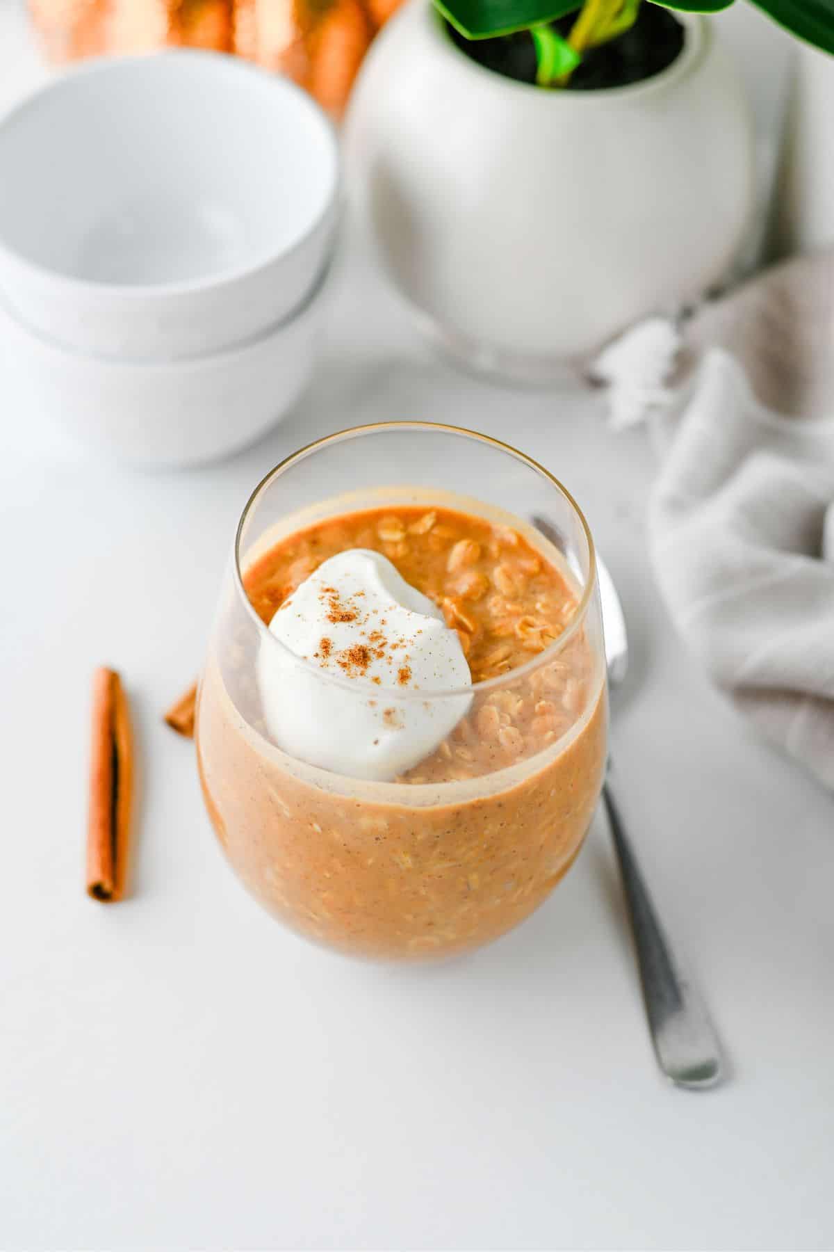 A glass of pumpkin-colored Pumpkin Overnight Oats topped with a dollop of whipped cream and sprinkled with cinnamon, placed on a white surface next to a spoon, a cinnamon stick, stacked bowls, and a potted plant.