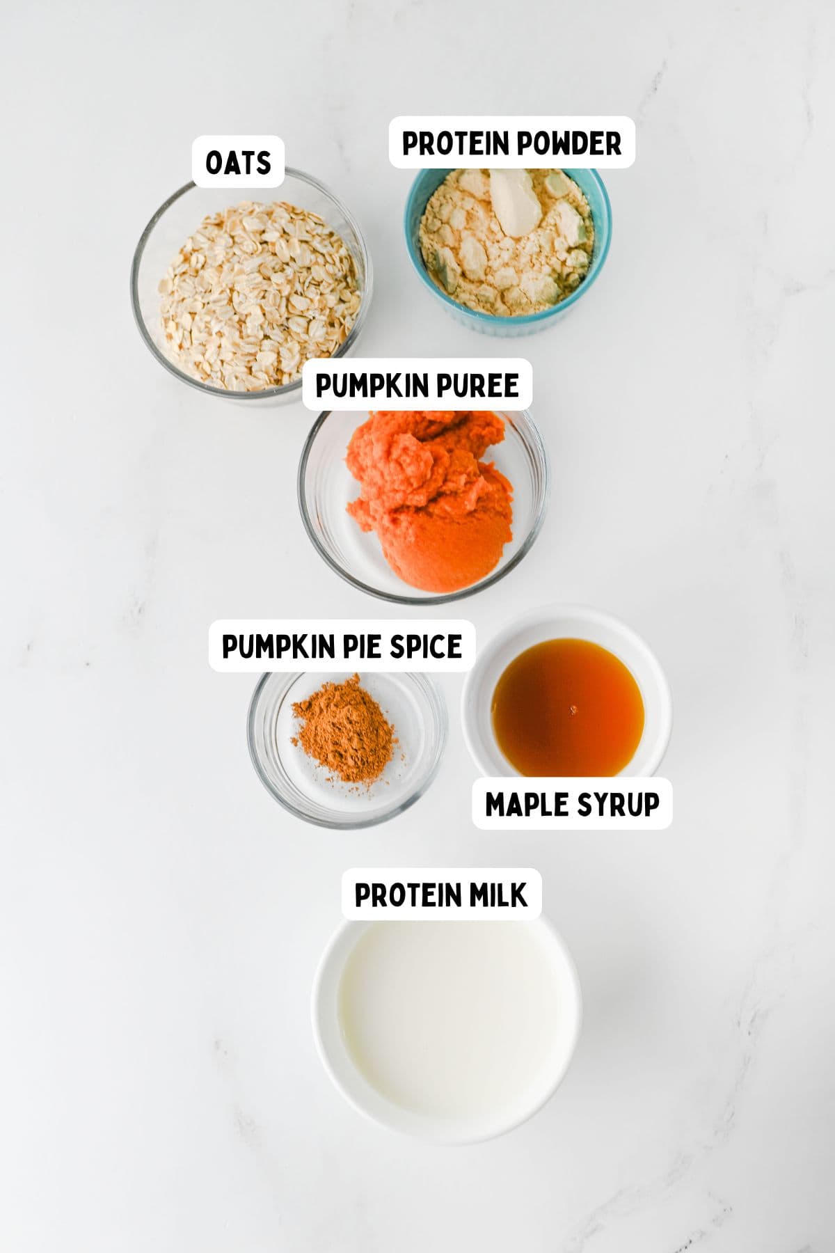 Six bowls on a white surface, each containing a labeled ingredient for Pumpkin Overnight Oats: oats, protein powder, pumpkin puree, pumpkin pie spice, maple syrup, and protein milk.