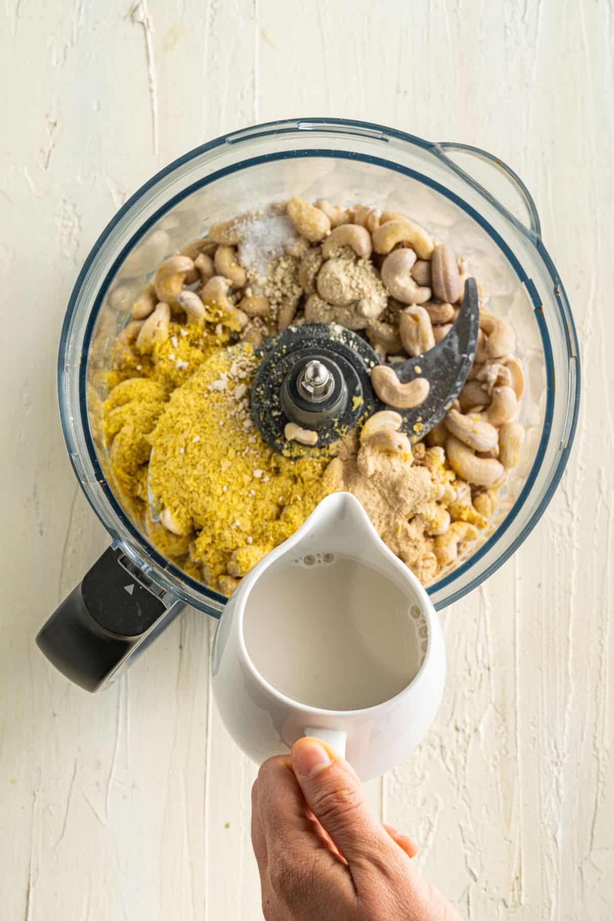 A food processor contains raw cashews, nutritional yeast, salt, and seasonings. A hand holds a white pitcher, pouring liquid—likely water or milk—into the processor. The items are on a light-colored wooden surface.