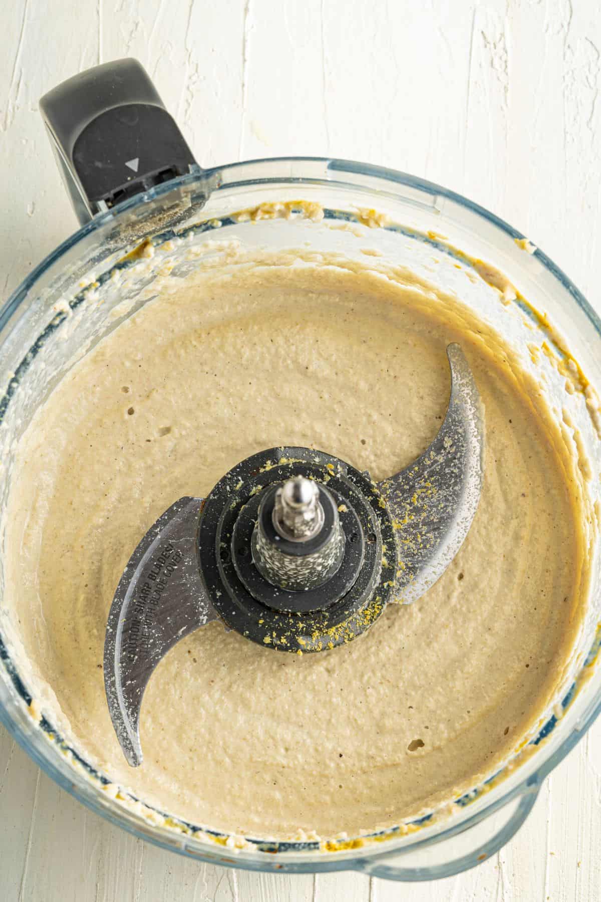 A food processor bowl containing a smooth, beige mixture, likely a batter or sauce, with some residue on the blade and sides. The processor is on a white, textured surface.