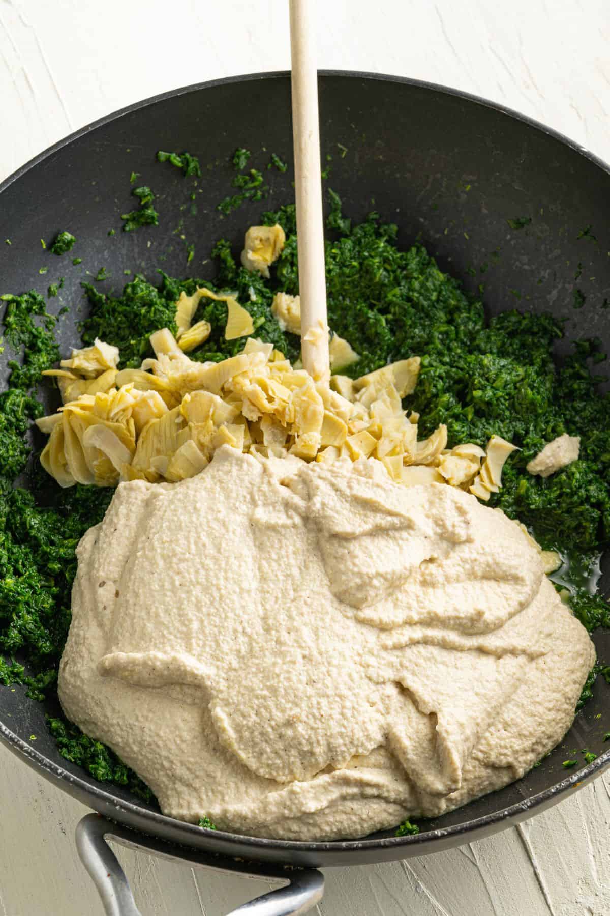 A skillet contains chopped spinach, chopped artichoke hearts, and a large portion of creamy mixture, being stirred together with a wooden spoon. The ingredients are not yet fully mixed.