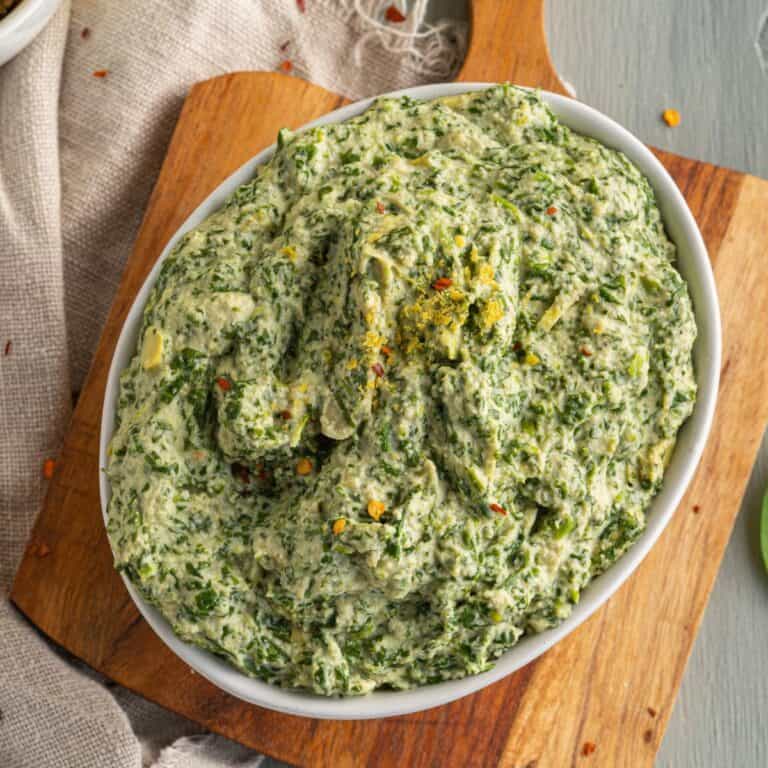 A white oval bowl filled with a creamy spinach dip, garnished with lemon zest and red pepper flakes, sits on a wooden board with a neutral cloth underneath.