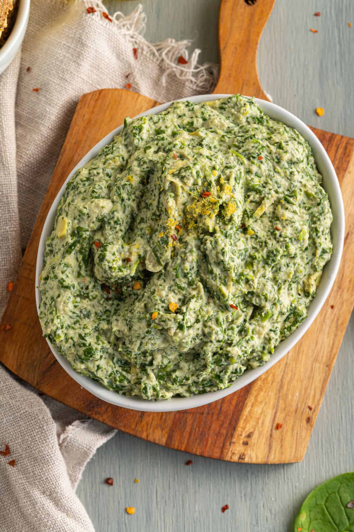 A white oval dish filled with creamy spinach artichoke dip sits on a wooden cutting board, with a beige cloth partially visible underneath. The dip is thick and speckled with herbs and red pepper flakes.