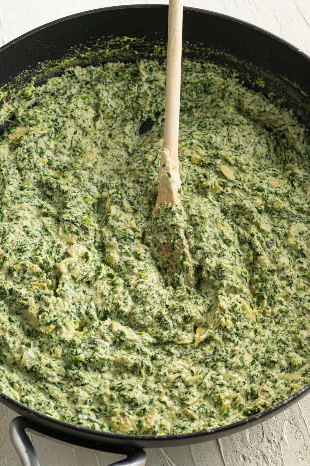A skillet filled with a creamy spinach and artichoke mixture being stirred with a wooden spoon, set on a light-colored surface.