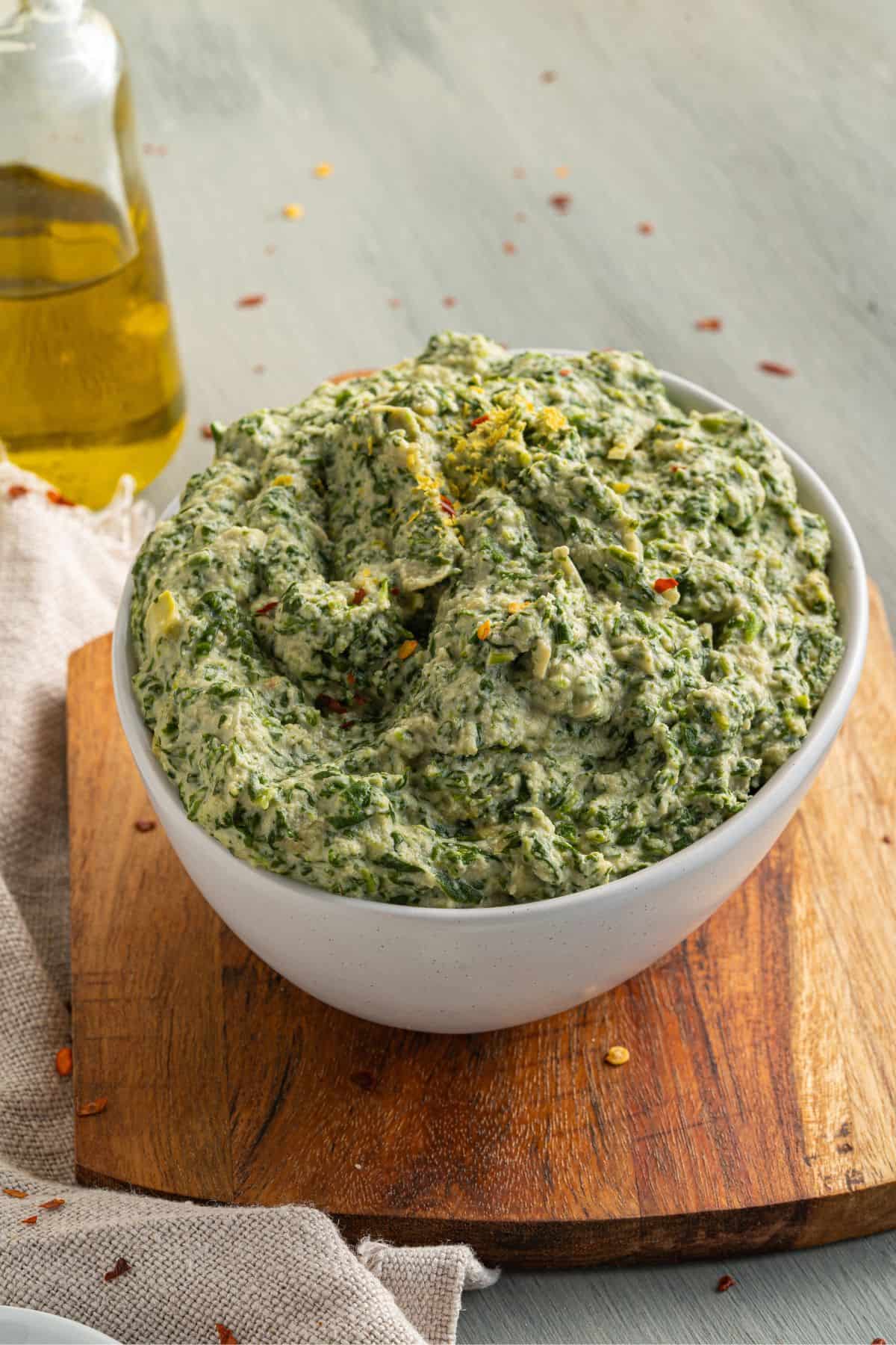 A white bowl filled with creamy spinach dip sits on a wooden board. A bottle of olive oil and a beige cloth are nearby on a light gray surface. Some dip is sprinkled with red pepper flakes.