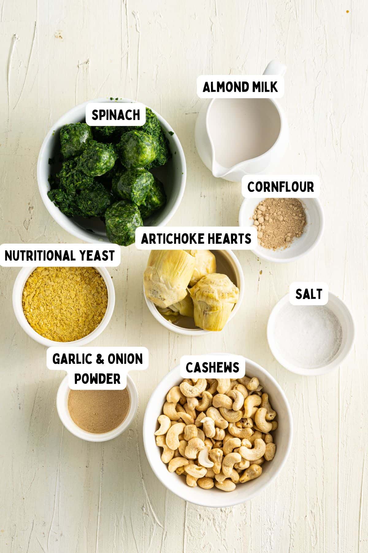 Eight small bowls on a light surface contain spinach, almond milk, cornflour, nutritional yeast, artichoke hearts, salt, garlic and onion powder, and cashews. Each bowl is labeled with its respective ingredient.