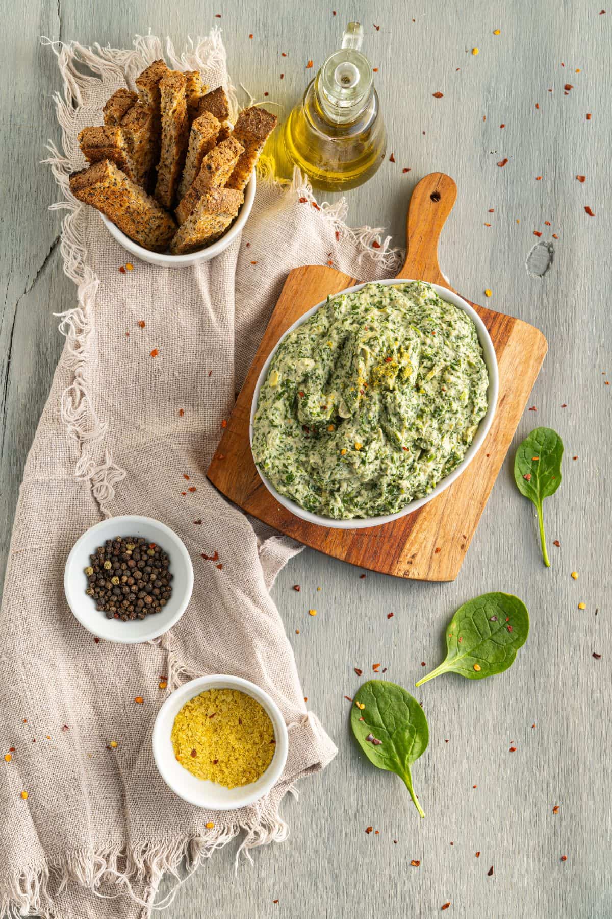 A bowl of spinach dip on a wooden board, surrounded by a bowl of toasted bread slices, a bottle of olive oil, a bowl of cracked pepper, a bowl of grated cheese, and spinach leaves on a light gray table.