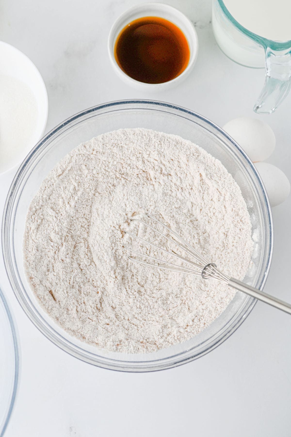 A wire whisk rests in a glass bowl filled with flour, surrounded by small containers of sugar, vanilla extract, milk, and eggs.