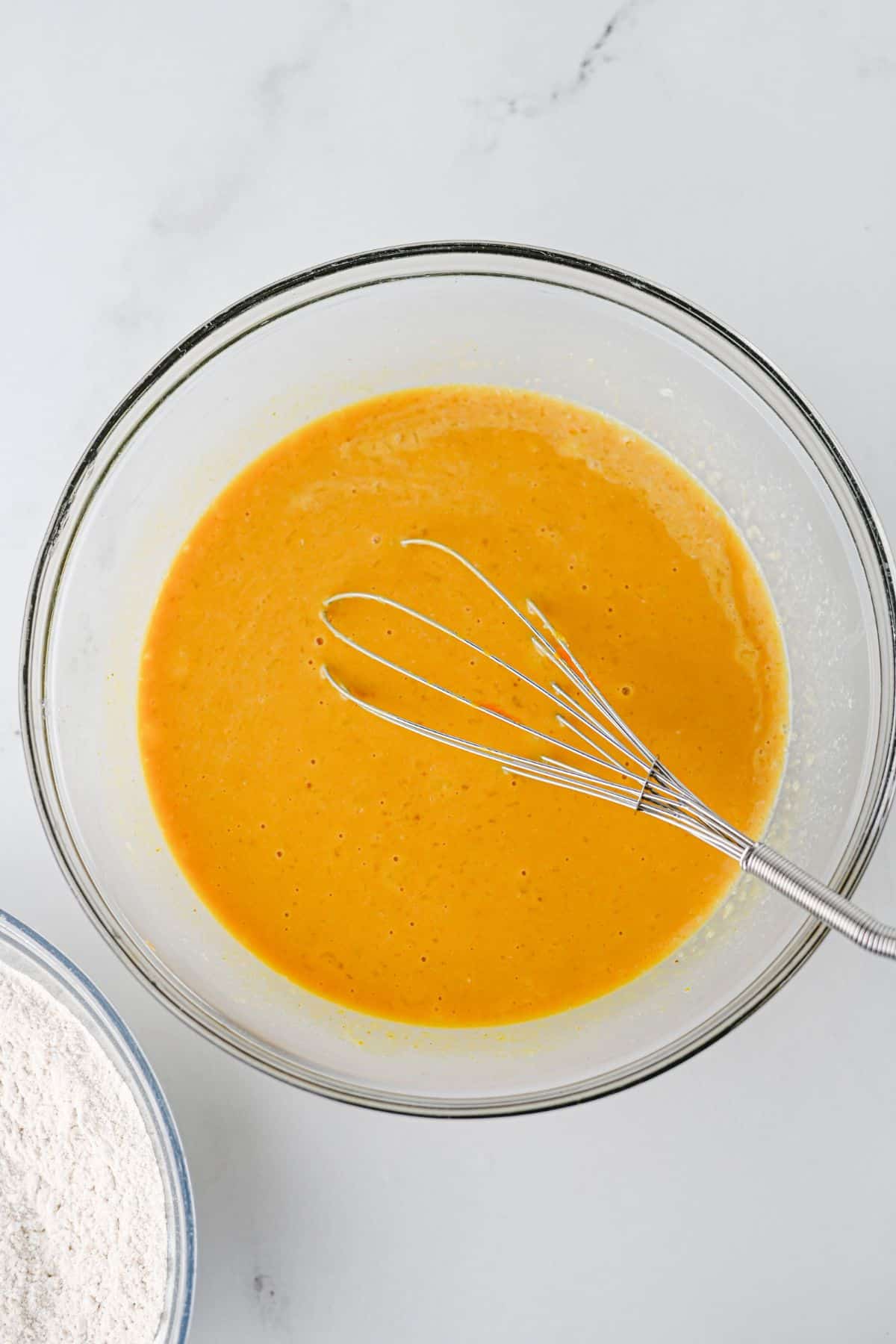 A glass bowl filled with orange batter and a metal whisk, with a corner of a bowl of flour visible.